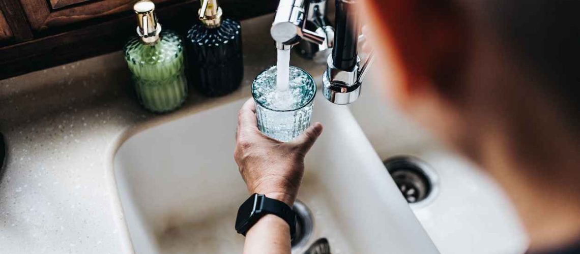 Over the shoulder view of a man filling a glass of filtered water right from the tap in the kitchen at home